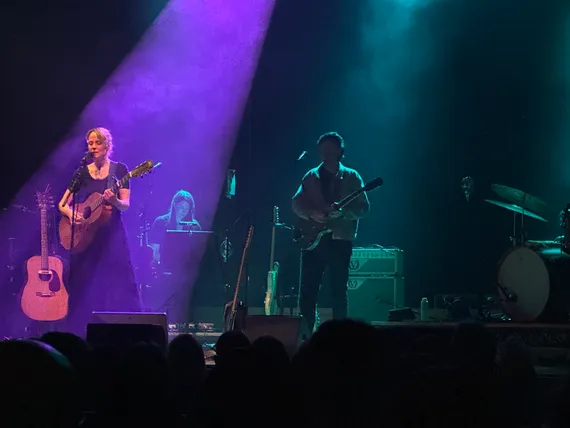 A blonde woman with an acoustic guitar under purple stage lights, with band members in shadow.