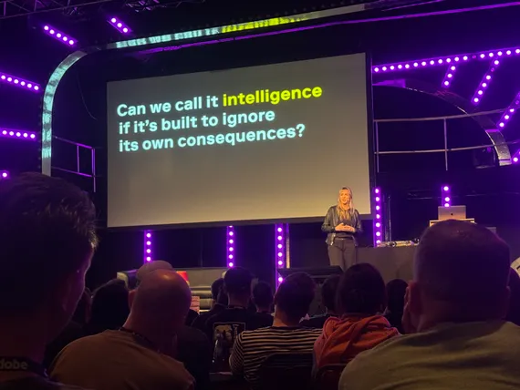 Lauren Celenza on stage in front of a slide reading 'Can we call it intelligence if it's built to ignore its own consequences?'