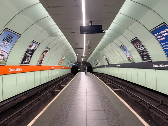 A subway station. Signage indicates the station is named Cowcaddens. The curve of the walls mimics the two subway tunnels in the background. One of the ads that line the walls says 'Hello Glasgow.' A single passenger approaches from the other end of the platform.