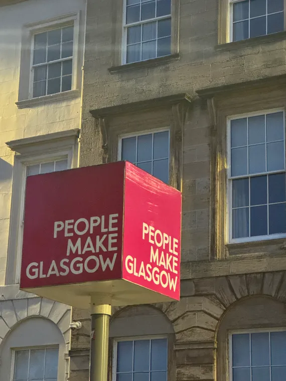 A pink sign reads 'People Make Glasgow', in front of a sandstone townhouse.