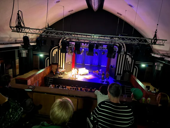 High angle photo of a stage, with four actors on a simple living room set, and a musical accompanist.