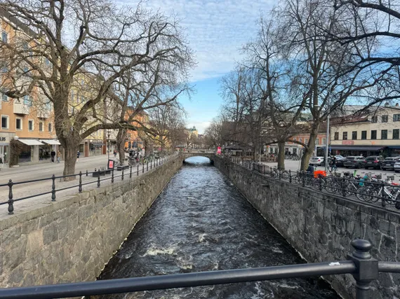 A river running between stone walls, with streets on either side and a stone bridge in the distance.